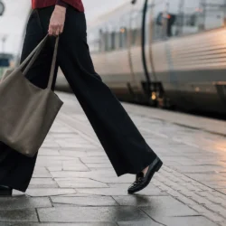 Vide sorte arbejdsbukser over sorte loafers på en togperron