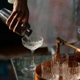Cocktailshaker der hælder en drink i et coupeglas ved hjemmebaren