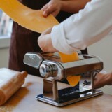 Hænder der kører pastadej gennem en manuel pastamaskine i et køkken