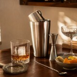 Cocktailshaker, jigger, si og cocktailglas på et varmt træbord i en hjemmebar