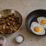 Stegepande i rustfrit stål med brunede svampe ved siden af nonstickpande med spejlæg
