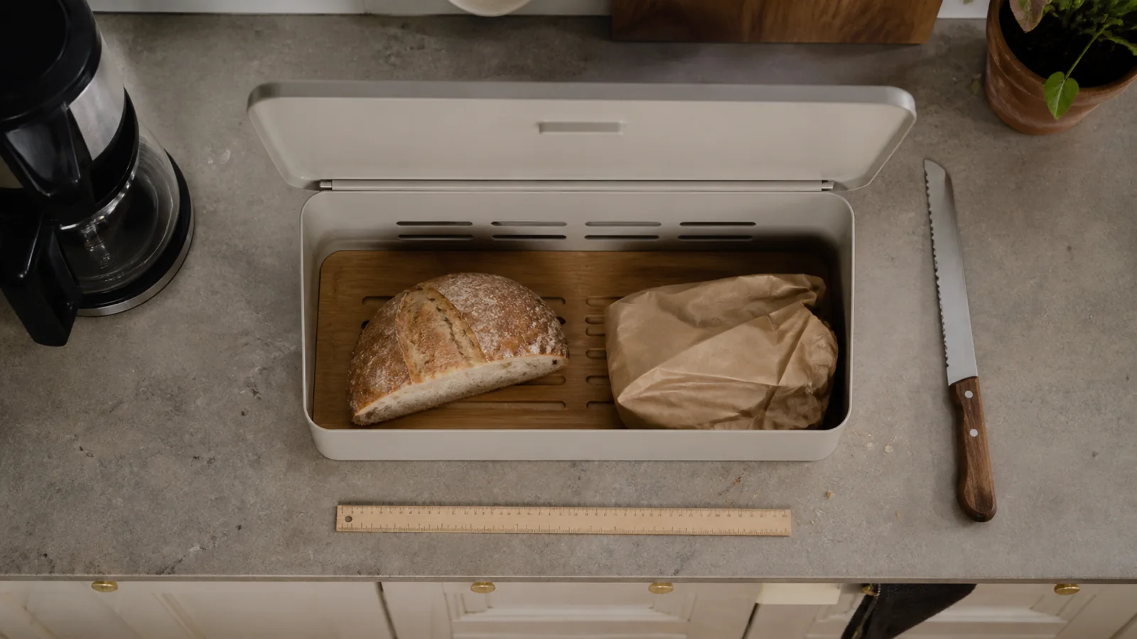 Åben brødkasse på køkkenbord med lineal, brødkniv og kaffemaskine omkring sig