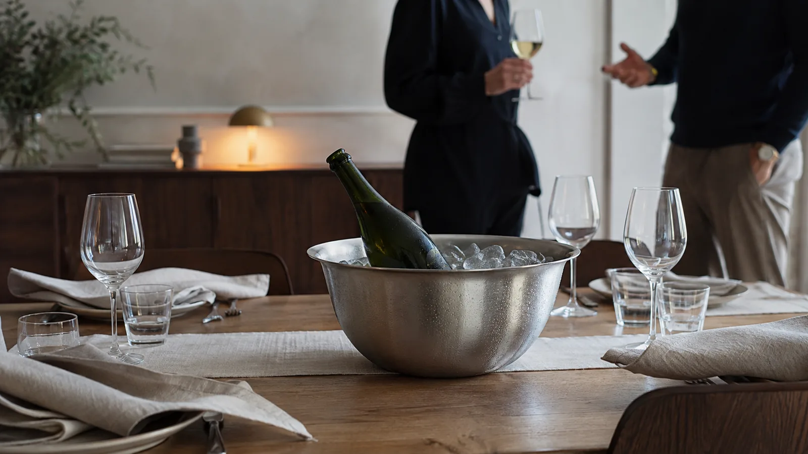 Champagneflaske i en stålskål med is og vand på et dækket bord, mens gæsterne ankommer