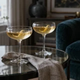 To coupeglas med lyse cocktails på et glasbord ved en petrolfarvet lænestol