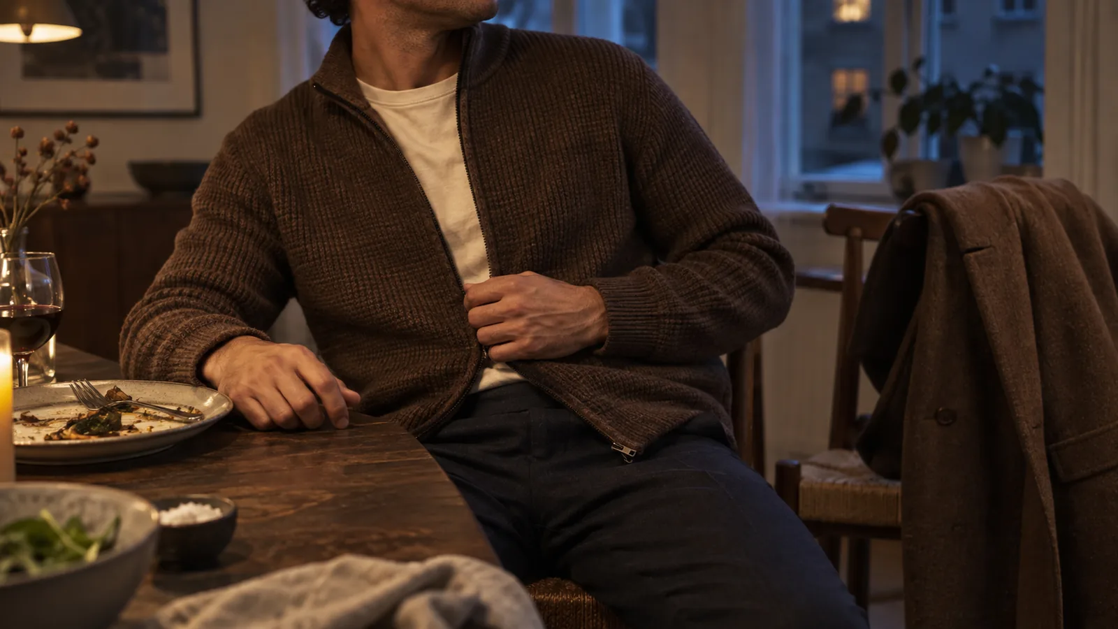 Brun cardigan over råhvid T-shirt ved middagsbordet, mens frakken hænger på stolen